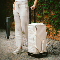 Person pulling a white suitcase on a gravel path with greenery in the background.